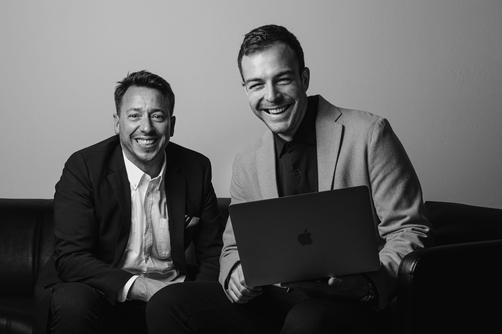 Benjamin Stöcklein und Matthias Räder, die Gründer der BeMa Group, lachen gemeinsam auf einem schwarzen Sofa vor heller Wand, einer hält einen aufgeklappten Laptop.
